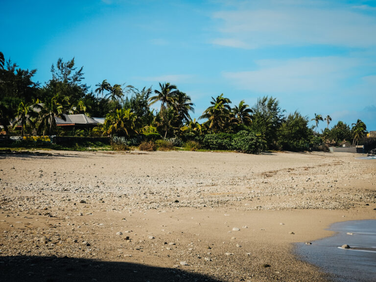 Szeroka, piaszczysta plaża z drobnymi kamieniami, za którą ciągnie się gęsty pas palm kokosowych i agaw skrywający domki z szarymi dachami pod bezchmurnym niebem.