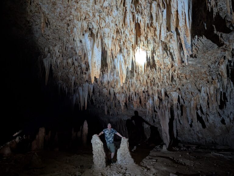 Turysta stoi pośród gigantycznych formacji stalaktytów i stalagmitów w ciemnym wnętrzu jaskini Hoq, oświetlonym punktowym światłem latarki.
