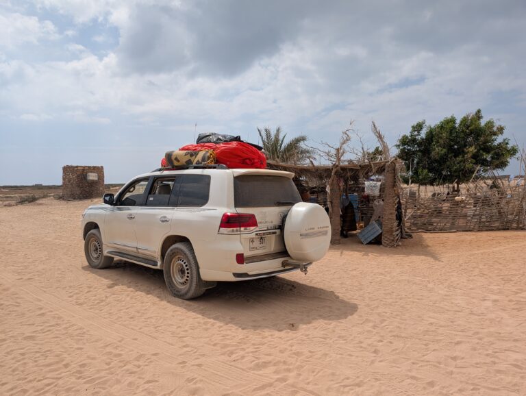 Biała Toyota Land Cruiser z bagażem wyprawowym na dachu, zaparkowana na piaszczystym terenie obok tradycyjnej wiaty z liści palmowych i kamiennej wieży.