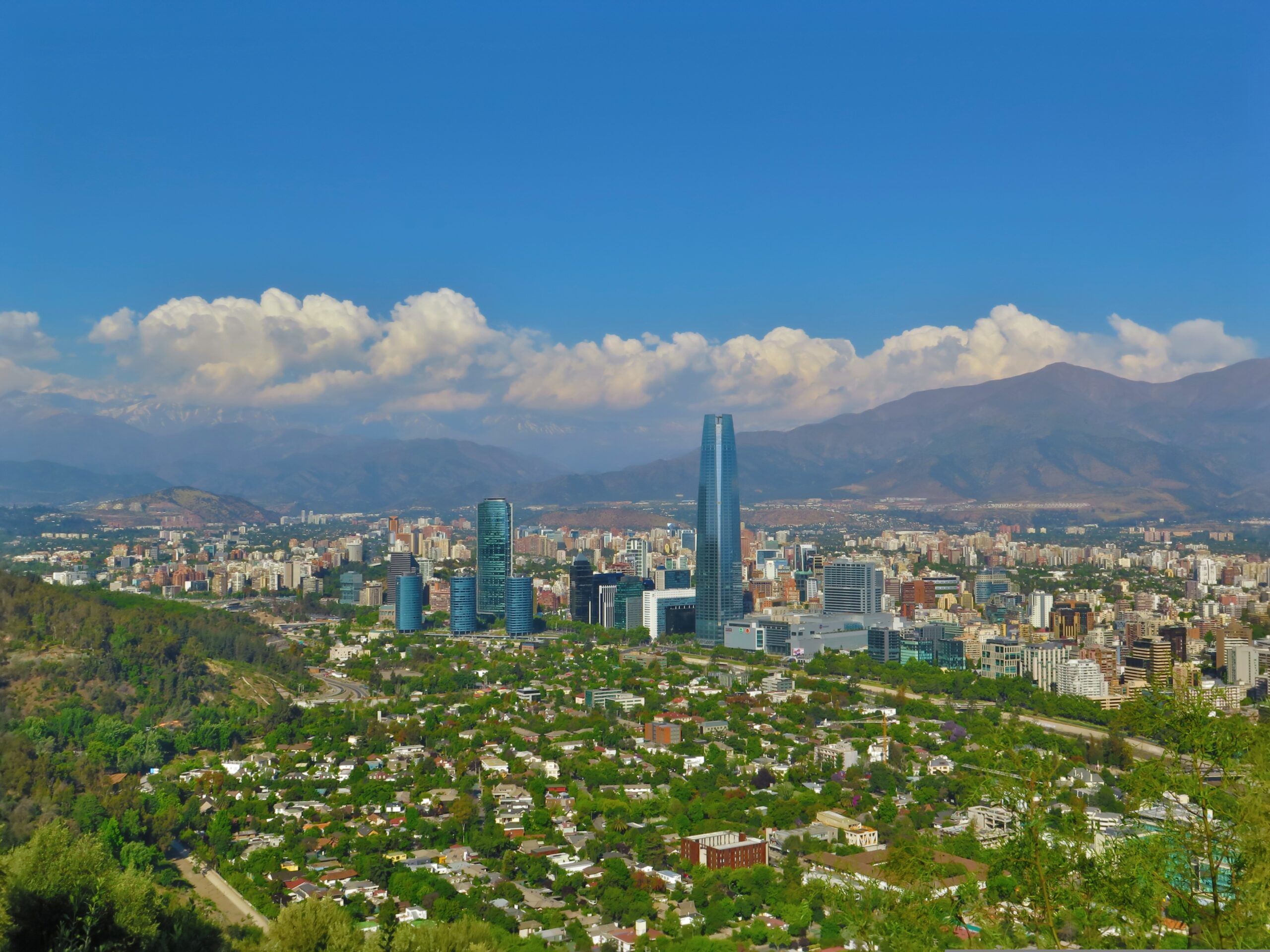Szeroki widok na panoramę Santiago z dominującym wieżowcem Gran Torre Santiago, otoczonym zielenią podmiejskich osiedli, z majestatycznymi szczytami Andów w tle pod bezchmurnym niebem.