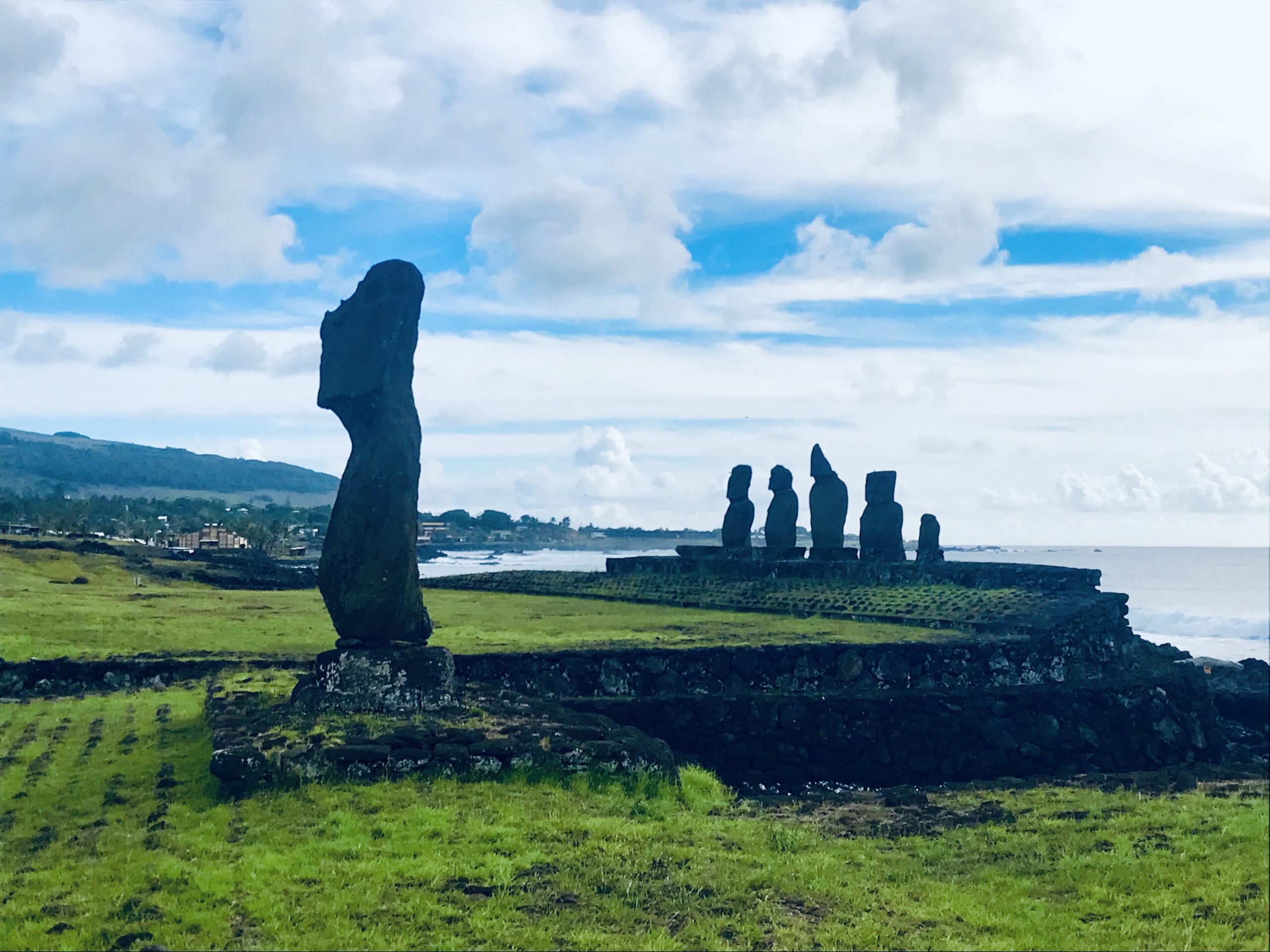Sylwetka samotnego moai na pierwszym planie oraz grupa posągów w oddali na tle zielonych pól i wzburzonego oceanu na Rapa Nui.