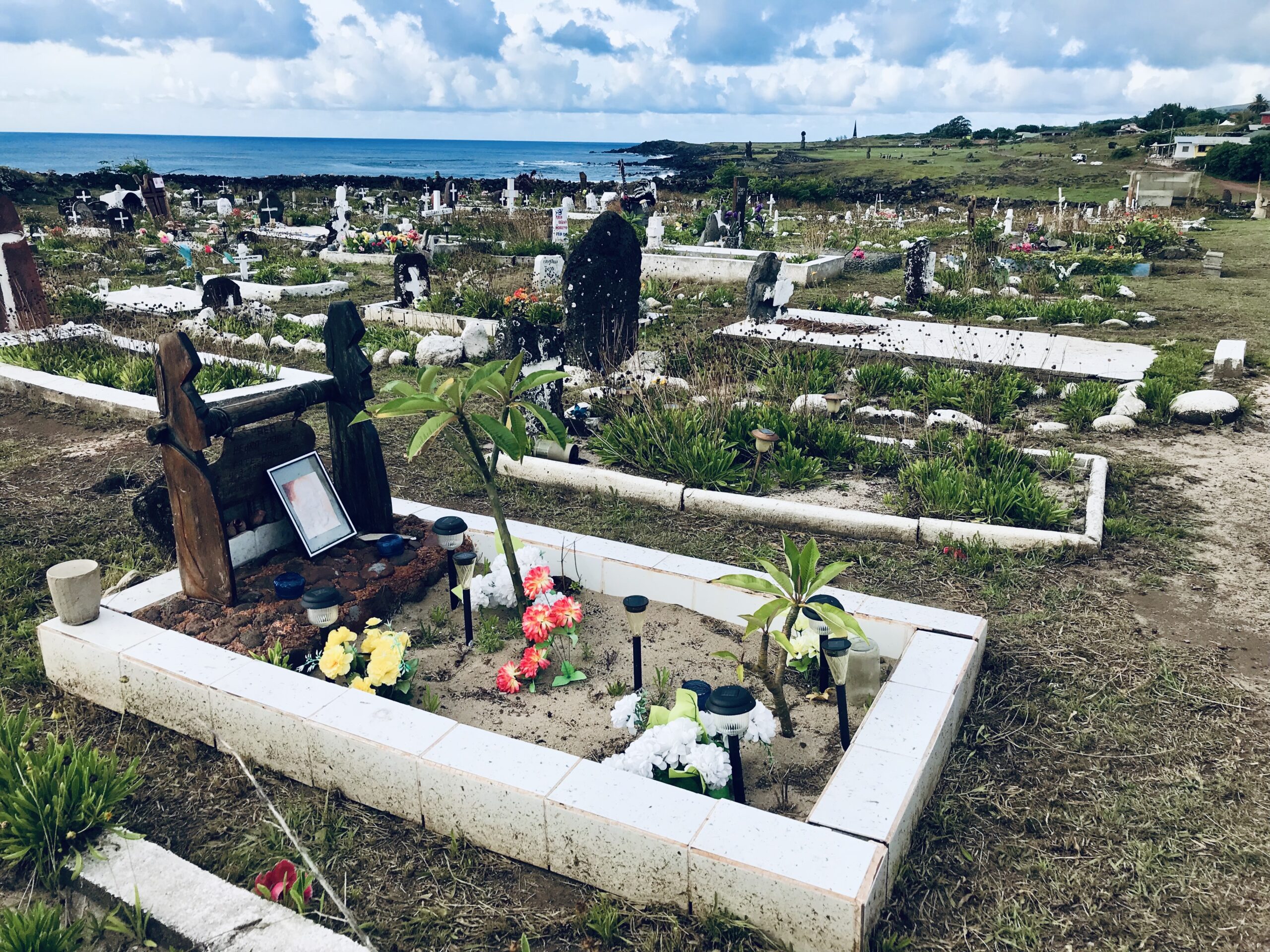 Panorama lokalnego cmentarza na Wyspie Wielkanocnej z białymi grobami ozdobionymi kwiatami, położonego tuż nad brzegiem błękitnego oceanu.